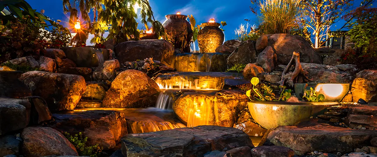 Backyard waterfall with illuminated rocks at night, showcasing water feature landscaping design and outdoor lighting.