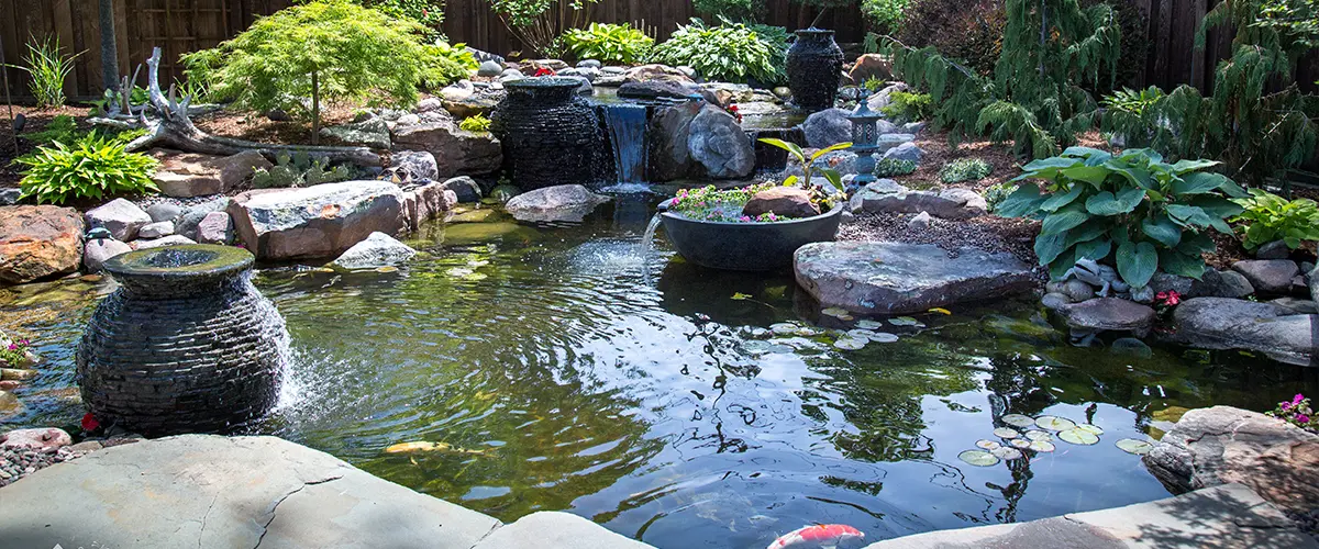 Beautiful backyard pond with waterfall surrounded by lush greenery and rock formations, creating a serene outdoor oasis.