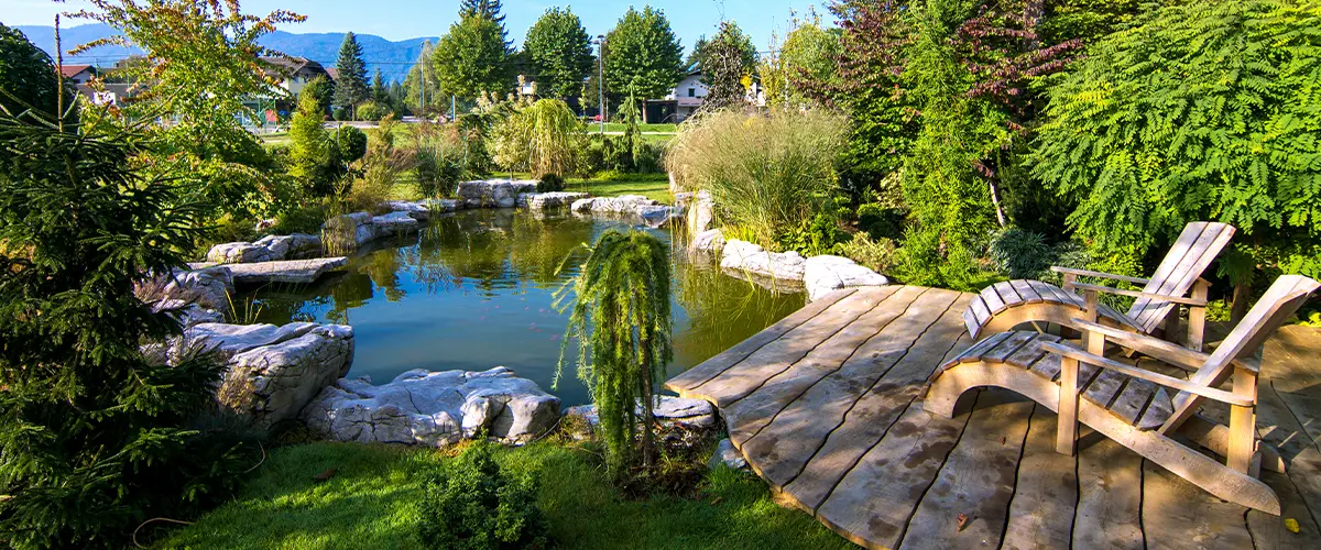 Idyllic backyard with a pond, lush greenery, and a wooden deck featuring lounge chairs, perfect for relaxation in a natural setting.