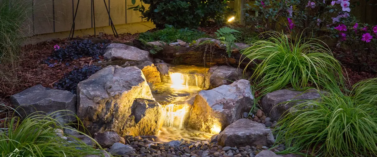 Small illuminated waterfall in a backyard garden with rocks, flowers, and landscape lighting for nighttime ambiance.