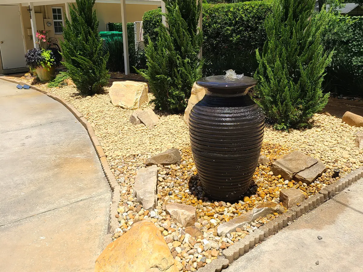 Stone urn fountain with pebble landscaping near driveway and shrubs