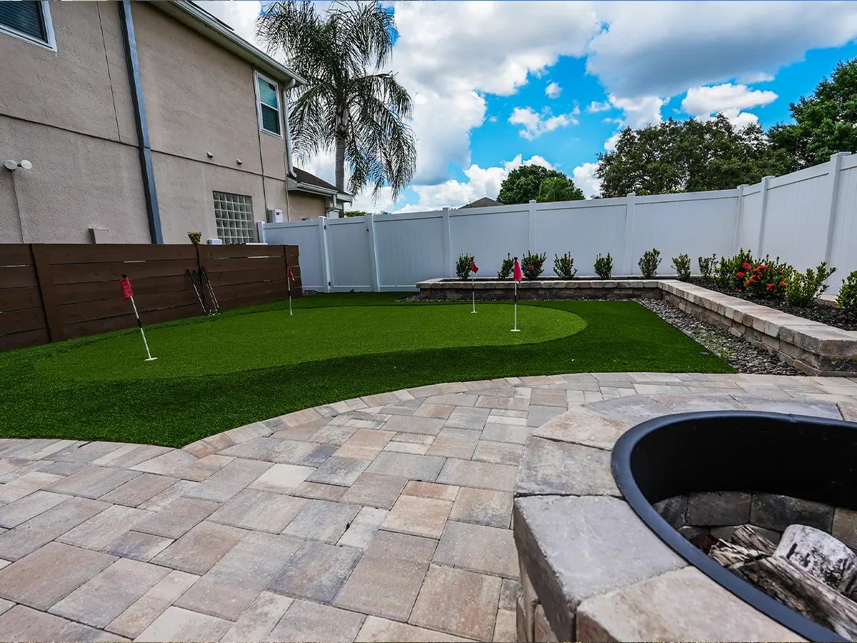 Transformed backyard with artificial turf putting green, modern patio, and fire pit, showcasing luxury landscaping design.