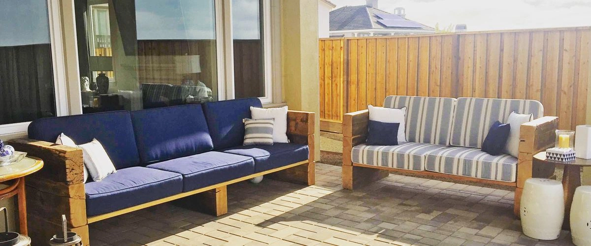 Spacious backyard paver patio furnished with two modern wooden-frame sofas with blue cushions, against a wooden fence